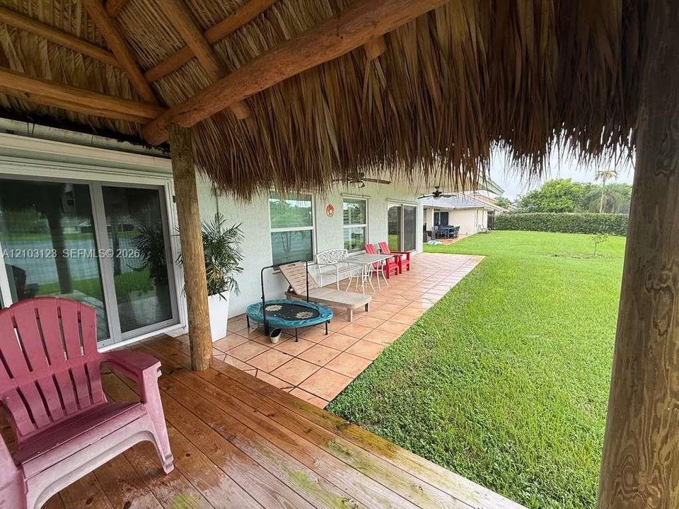 Covered patio area featuring a thatched roof, rustic timber support beams, and ceramic tile flooring, extending to a manicured lawn
