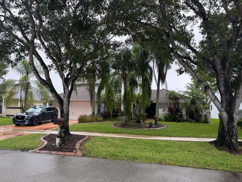 Exterior facade featuring a paved driveway, mature trees, manicured lawn, and professional landscaping
