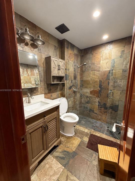 Bathroom featuring natural stone tile flooring and wall accents, a vanity with a white countertop and rectangular undermount sink, a wall-mounted cabinet, and a walk-in shower with a glass enclosure