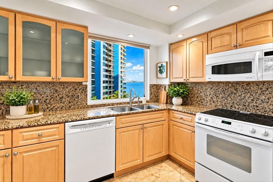 Kitchen featuring natural wood cabinetry with glass-front uppers, stone countertops, full-height stone backsplash, double-basin stainless steel sink, and white appliances