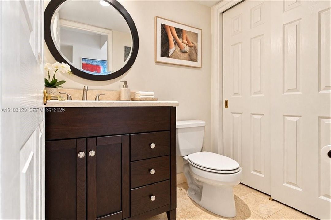 Dark wood vanity with a stone countertop, oval wall-mounted mirror, neutral stone flooring, and white two-panel closet doors