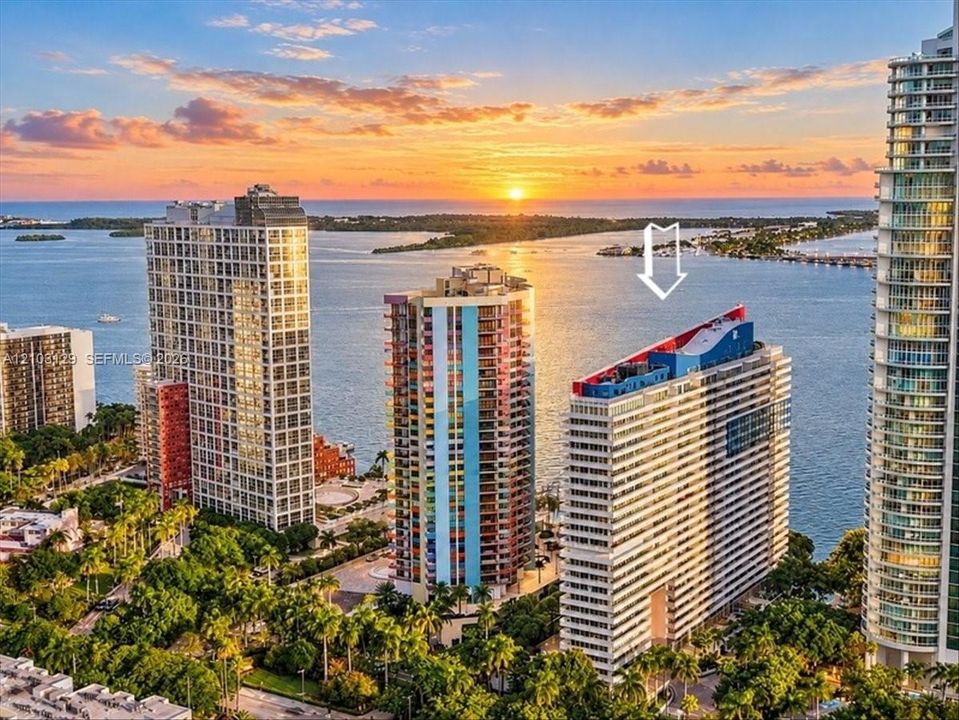 High-rise residential building featuring expansive coastal water frontage, architectural rooftop detailing, and surrounding mature tropical landscaping