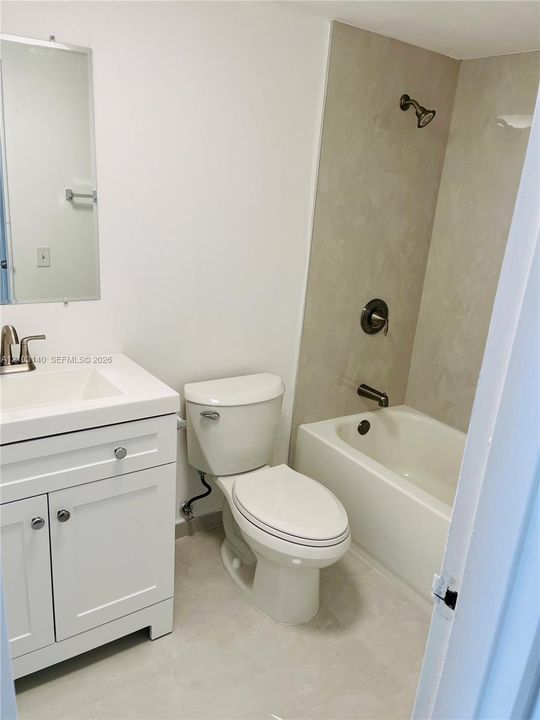 White vanity with a rectangular sink, wall-mounted mirror, two-piece toilet, and tub-shower combination featuring neutral wall surround and metallic fixtures