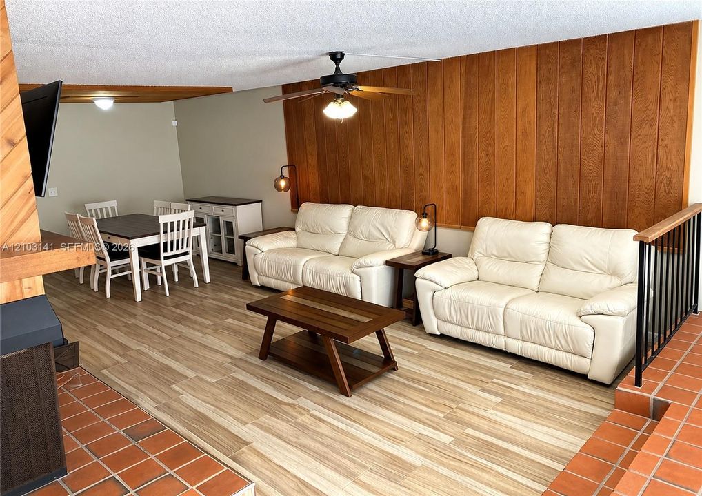 Wood-paneled accent wall featuring vertical grain, wood-finish flooring, terra-cotta tile hearth, black metal railing, and ceiling-mounted fan with integrated lighting