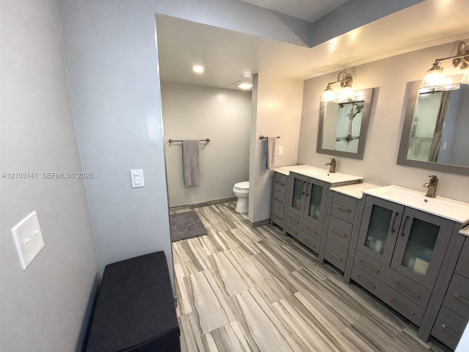 Double vanity featuring grey cabinetry with glass-panel doors, solid surface countertops, metallic finish fixtures, and wood-finish flooring
