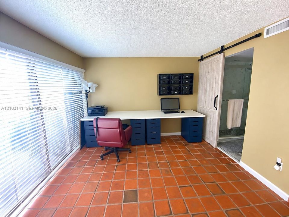 Terra-cotta tile flooring, large wall-to-wall window with horizontal blinds, built-in desk surface, and sliding wood-finish barn door