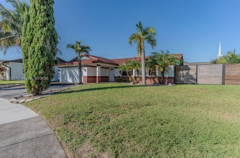 Single-story exterior featuring a brick facade, palm tree landscaping, a paved driveway, and horizontal wood-panel fencing