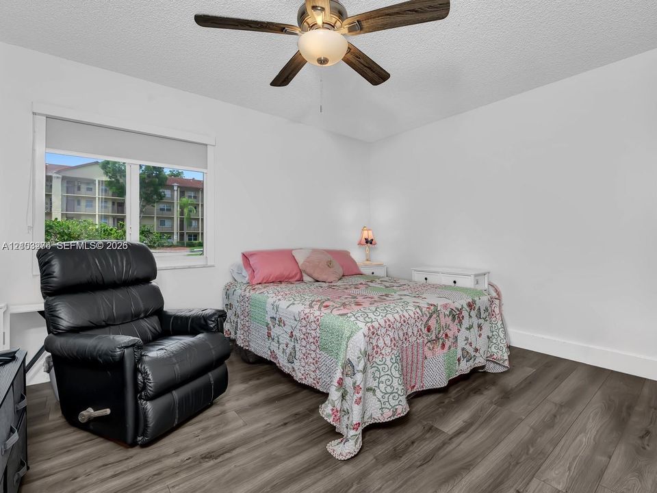 Room featuring wood-finish flooring, a multi-blade ceiling fan with integrated lighting, and a large window with a roller shade