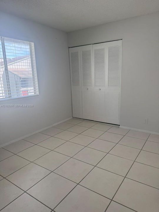Room featuring neutral tile flooring, window with white blinds, and built-in closet with louvered bi-fold doors