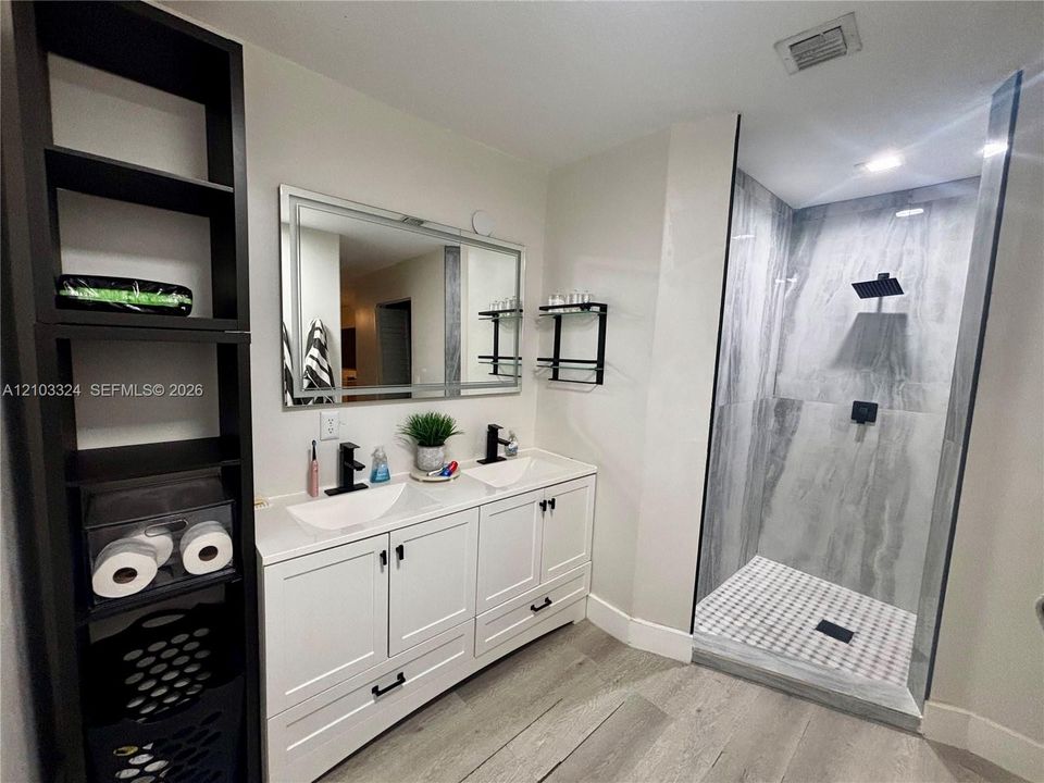 Dual-sink vanity with white cabinetry, rectangular vessel sinks, quartz countertops, and matte black fixtures, paired with a walk-in shower featuring large-format tile surround and wood-finish flooring