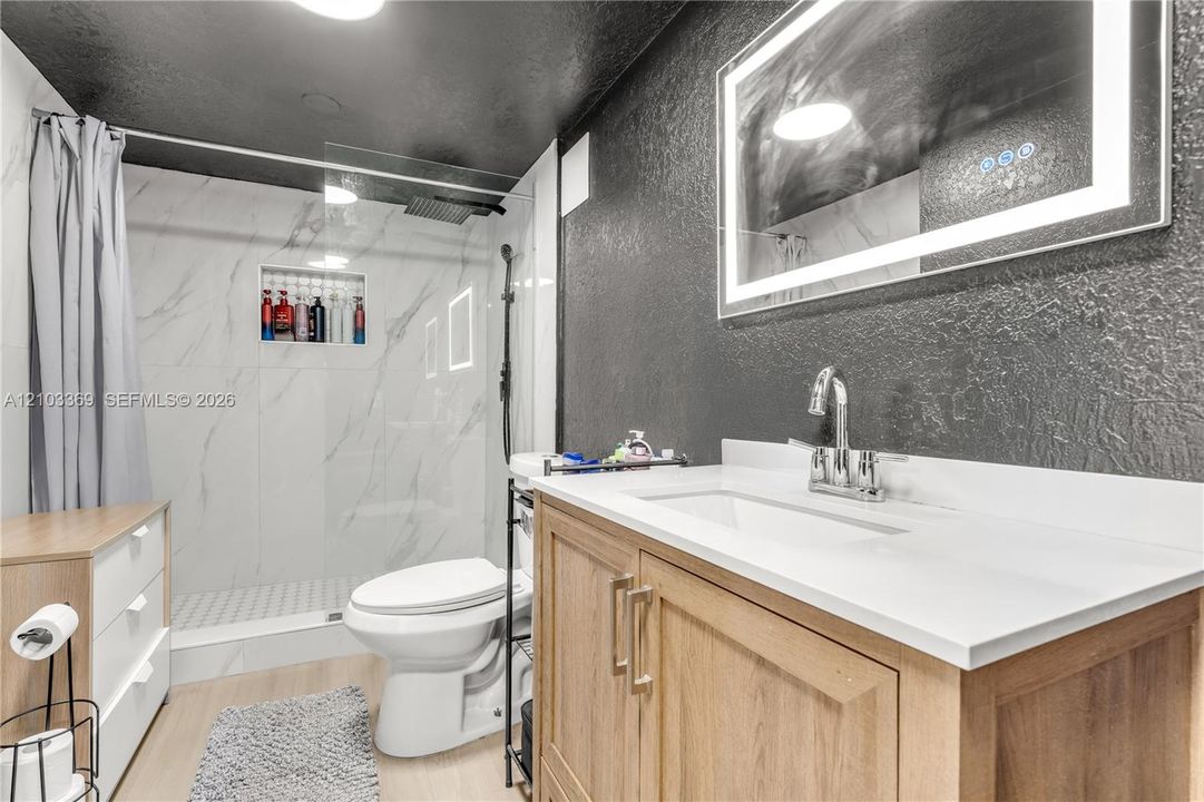 Bathroom featuring a wood-finish vanity with a stone-composite countertop, illuminated rectangular mirror, walk-in shower with floor-to-ceiling tile surround and built-in recessed shelving, and a rain-style showerhead