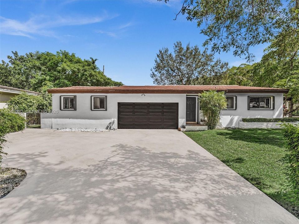 Single-story exterior featuring white stucco siding, a flat roofline, attached multi-car garage, concrete driveway, and manicured lawn. House for rent in Coral Gables. House for rent listed by Realtor