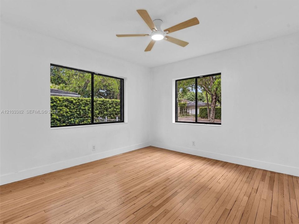 Large front/east bedroom with sunny windows. Featuring wood-finish flooring, two windows with black frames, and a multi-blade ceiling fan with integrated lighting. House for rent in Coral Gables. House for rent listed by Realtor.