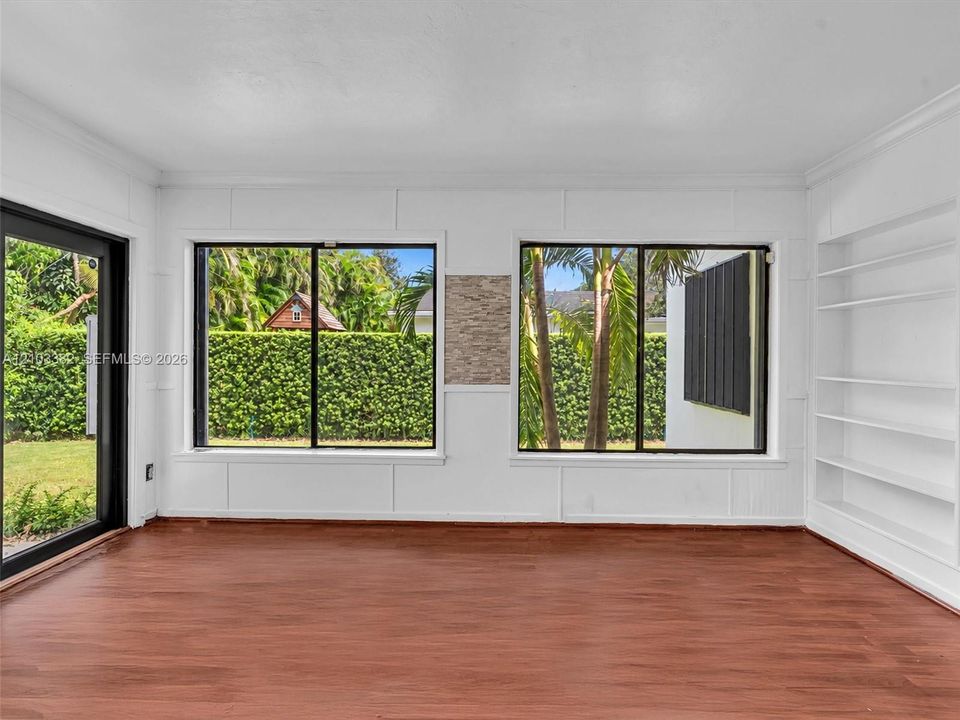 Florida room featuring wood-finish flooring, white panel-style wall detailing, large sliding glass doors, dual windows with black frames, and custom built-in shelving, and a large picture window. House for rent in Coral Gables. House for rent listed by Realtor.