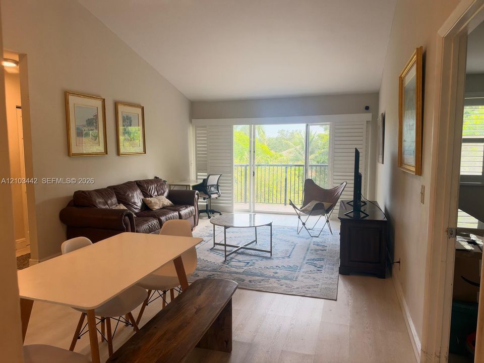Living area featuring vaulted ceilings, light-tone wood-finish flooring, and glass sliding doors with plantation shutters opening to a balcony with iron railings