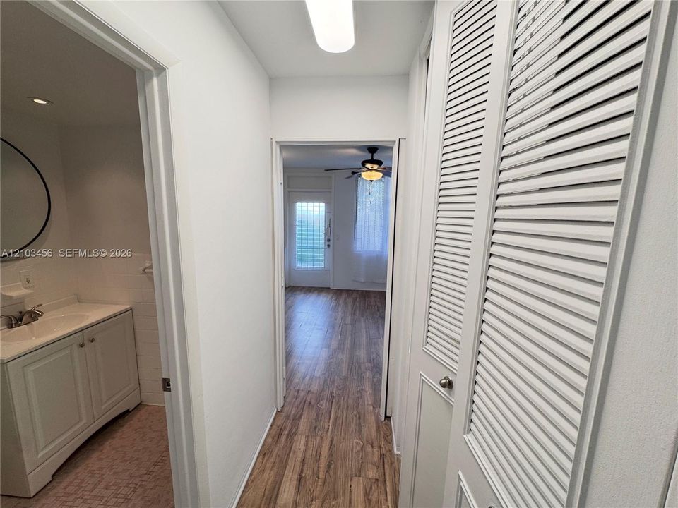 Hallway featuring wood-finish flooring, floor-to-ceiling bi-fold louvered closet doors, and integrated lighting, with a bathroom entry showcasing a vanity cabinet with a stone-top surface and a circular wall-mounted mirror