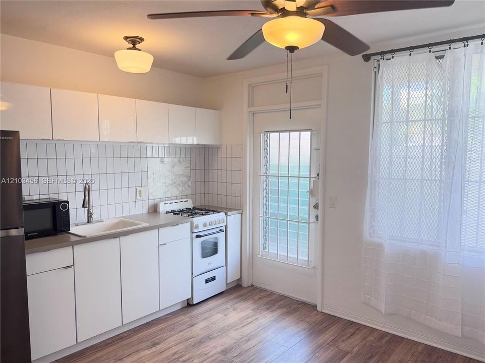 Kitchen featuring high-gloss white cabinetry, white square tile backsplash, wood-finish flooring, ceiling fan with light fixture, and exterior door with decorative security grate