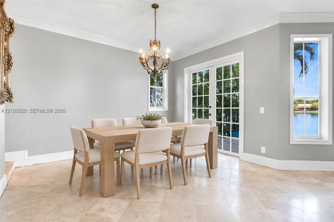 Neutral-toned interior space featuring light-colored stone flooring, intricate crown molding, a decorative metal chandelier, glass-paned French doors, and a multi-pane window with water scenery