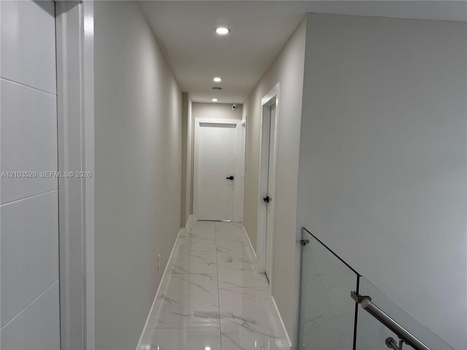 Hallway featuring large-format tile flooring with a neutral veined pattern, recessed ceiling lighting, glass panel railing with metallic hardware, and minimalist interior doors with matte black levers