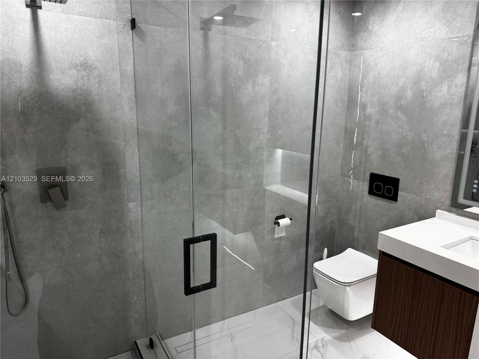 Walk-in shower with floor-to-ceiling stone-finish tile, glass enclosure, and wall-mounted plumbing fixtures, paired with a wall-hung toilet and floating vanity featuring a quartz countertop
