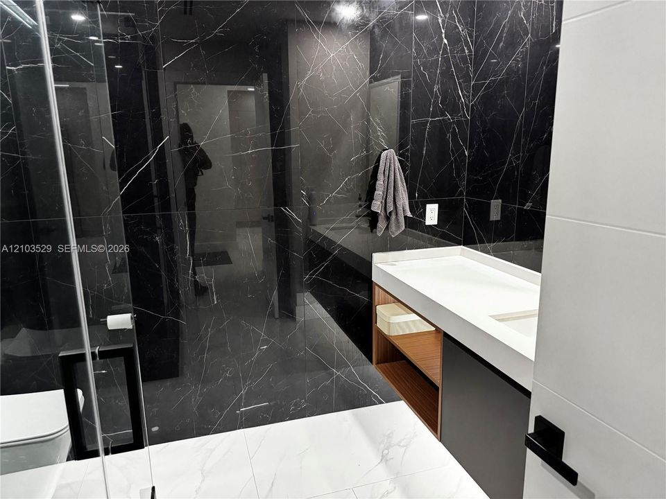 Modern bathroom featuring large-format dark veined stone wall tile, floating wood-finish vanity with a white countertop, glass-enclosed shower, and matte black hardware