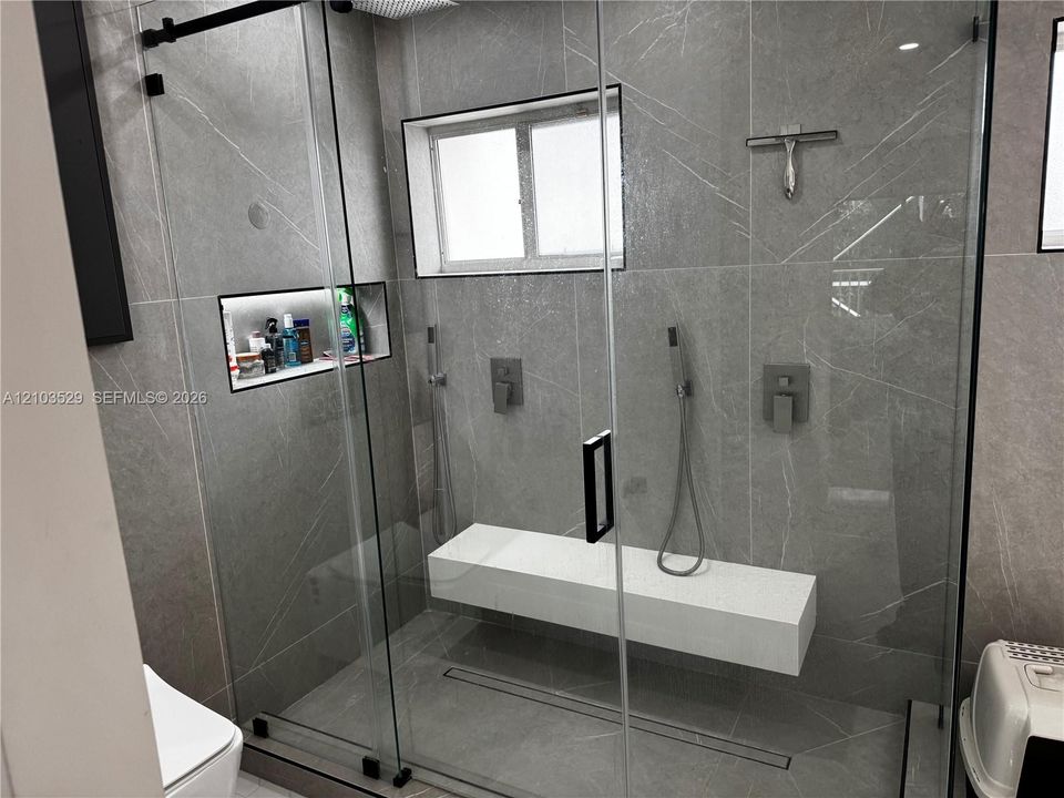 Walk-in shower featuring large-format tile walls, a sliding glass door, a floating stone bench, recessed wall shelving, and dual wall-mounted shower fixtures