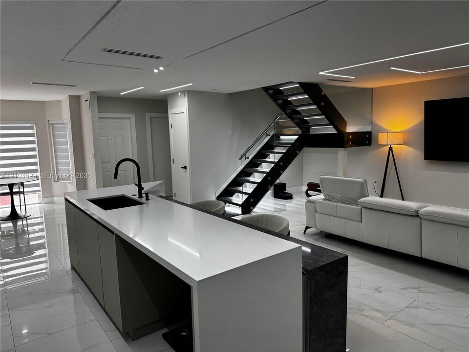Kitchen island featuring a white stone-surface countertop, matte black faucet, and an integrated dark stone-surface breakfast bar, complemented by glossy tile flooring, recessed linear lighting, and an open-tread staircase with glass railings