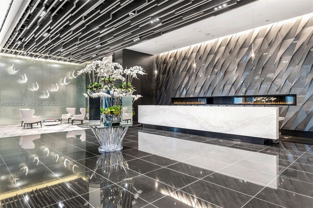 Reflective dark tile flooring, a marble-finish reception desk, a linear fireplace, an illuminated decorative wall panel, and a striking ceiling design