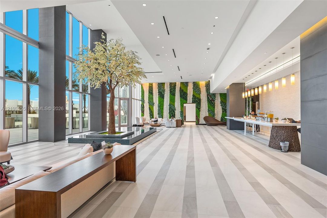 Expansive lobby with floor-to-ceiling windows, light-toned striped flooring, a built-in planters, a green wall feature, and recessed lighting