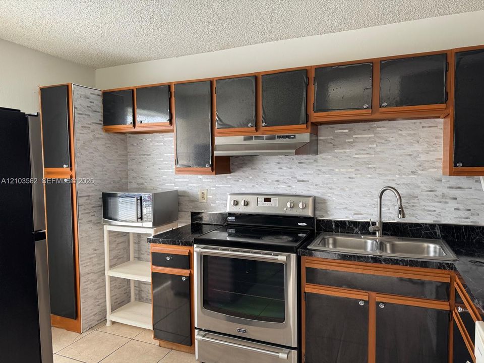 Kitchen featuring wood-trim cabinetry with black paneling, textured linear-tile backsplash, stainless steel electric range, and double-basin stainless steel sink with high-arc faucet