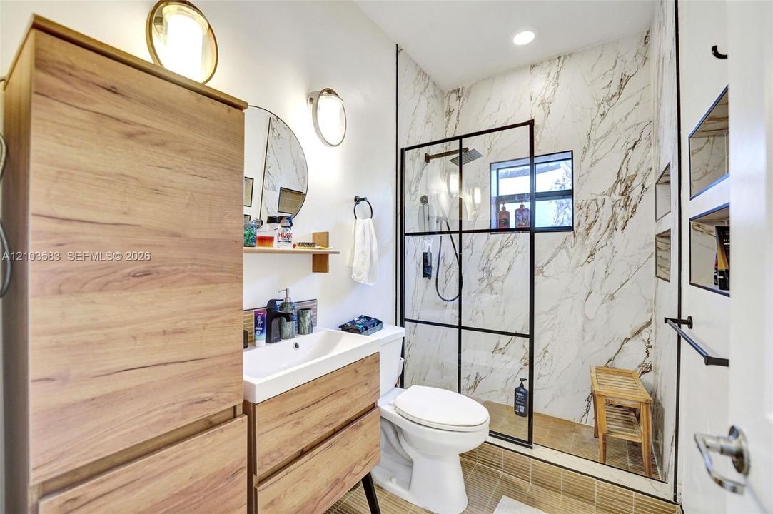 Bathroom featuring wood-finish cabinetry, a walk-in shower with black-frame glass enclosure and stone-pattern wall tile, circular vanity mirror, and wall-mounted light fixtures