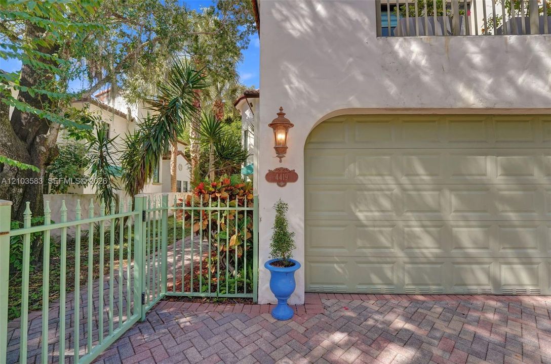 Stucco exterior featuring a paved brick driveway, metal gate enclosure, attached garage, and decorative wall-mounted lighting
