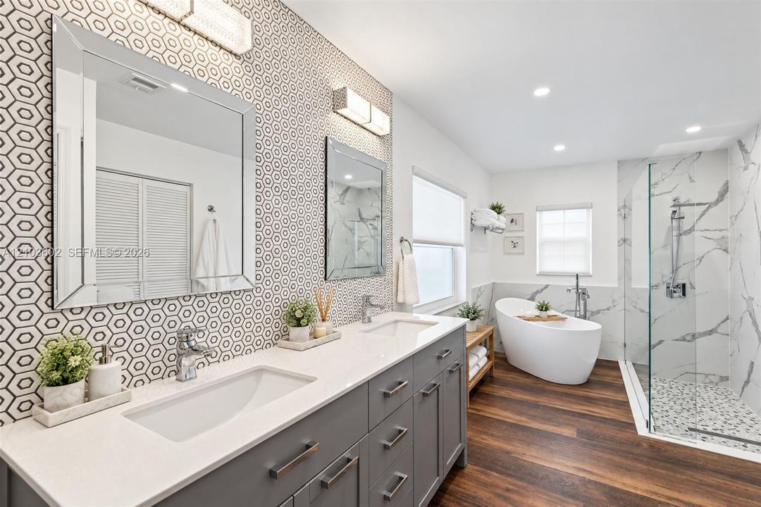 Bathroom featuring a double vanity with grey cabinetry, quartz countertops, a freestanding soaking tub, a walk-in glass-enclosed shower with stone tile flooring, and wood-finish flooring