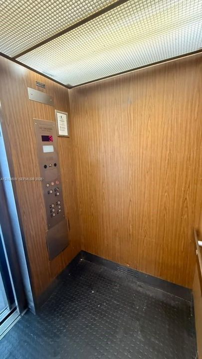 Elevator interior featuring wood-finish paneling, grid-patterned ceiling lighting, metallic control panel, and textured flooring