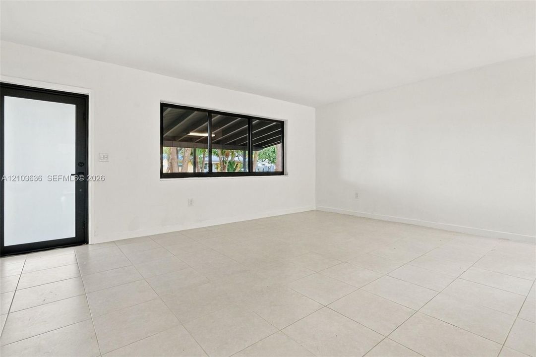Neutral interior featuring large-format tile flooring, stark white walls, a black-framed glass exterior door, and a multi-pane horizontal window