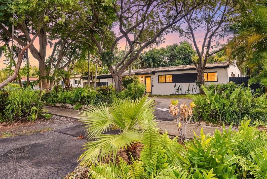 Single-story exterior featuring a light-toned stucco facade, dark-framed windows, a covered entryway, and mature shade trees within a lush, landscaped setting