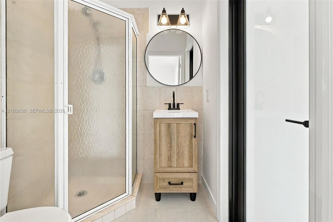 Corner shower enclosure with textured glass, wood-finish vanity featuring matte black hardware, circular wall-mounted mirror, and dual-bulb industrial-style lighting fixture