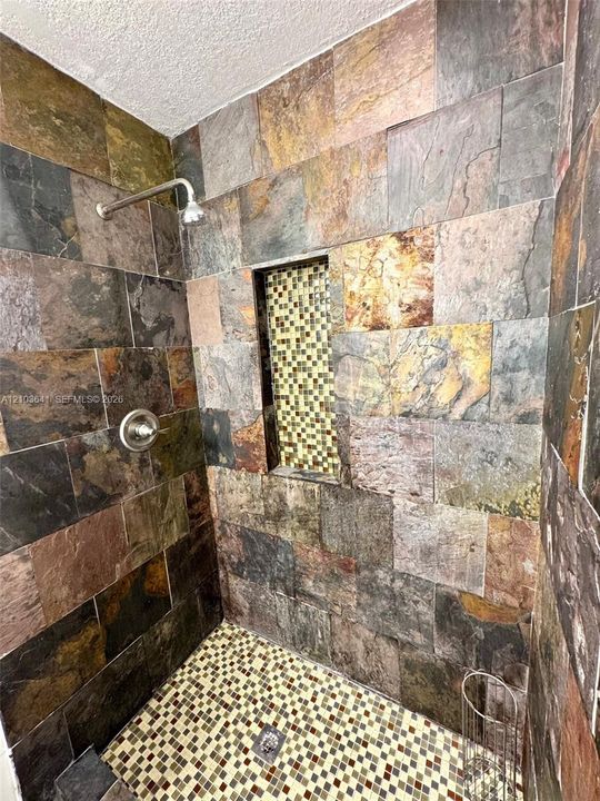 Walk-in shower featuring multi-toned natural stone wall tiles, a recessed tile-accented niche, mosaic floor tiling, and a metallic shower fixture