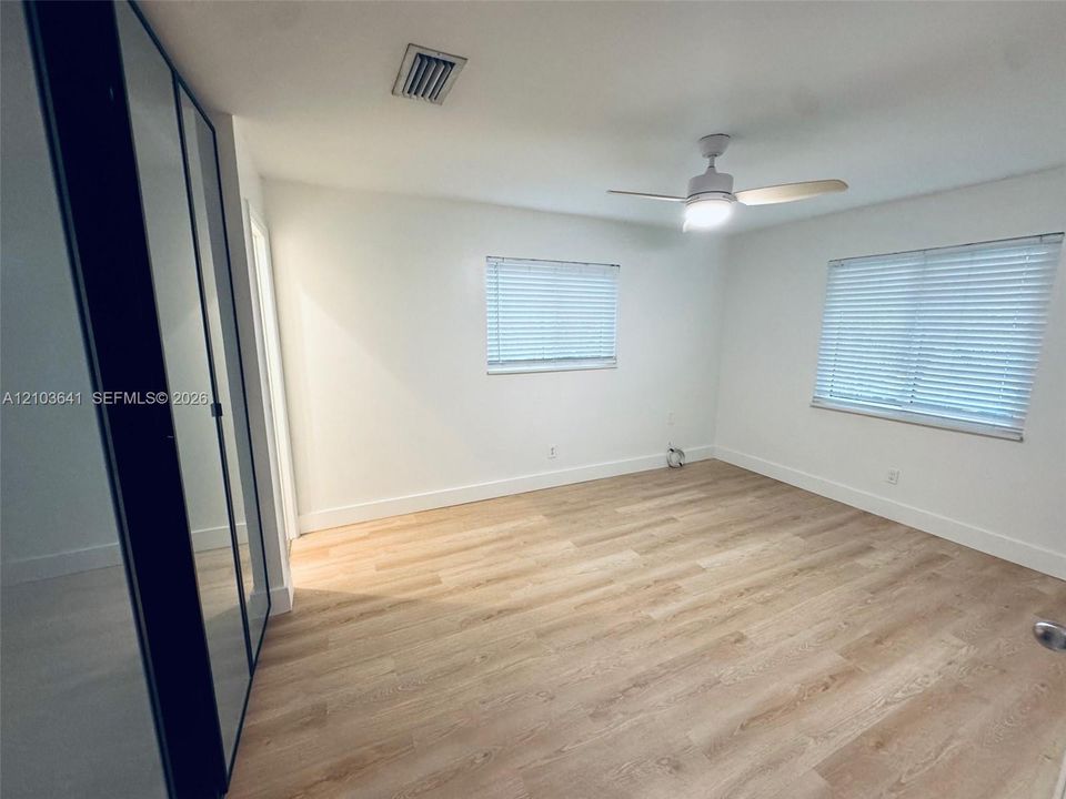 Light-filled room featuring wood-finish flooring, dual windows with blinds, a ceiling fan with integrated lighting, and full-length sliding mirror-paneled closet doors