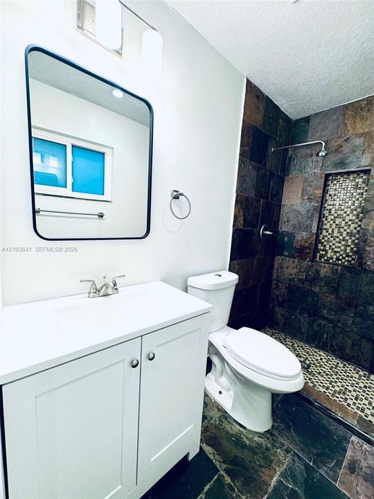 White vanity with integrated sink, rectangular mirror with black frame, multi-tone slate tile shower surround, mosaic tile shower inlay, and stone-finish floor tiling