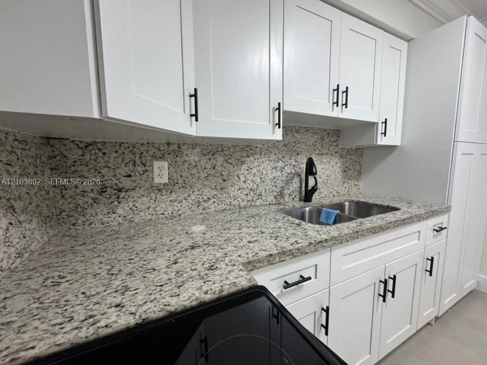 White Shaker-style cabinetry featuring matte black hardware, stone countertops with matching full-height backsplash, stainless steel double-basin undermount sink, and matte black high-arch faucet