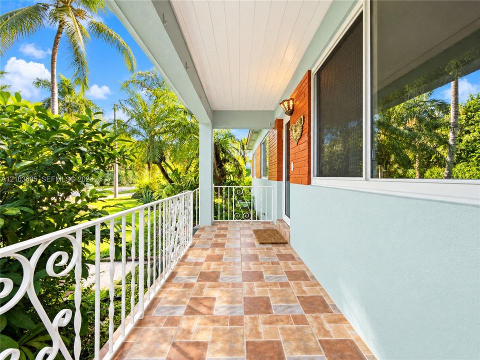 Covered porch featuring multi-tonal ceramic tile flooring, decorative wrought-iron railing, beadboard ceiling, and brick exterior accents