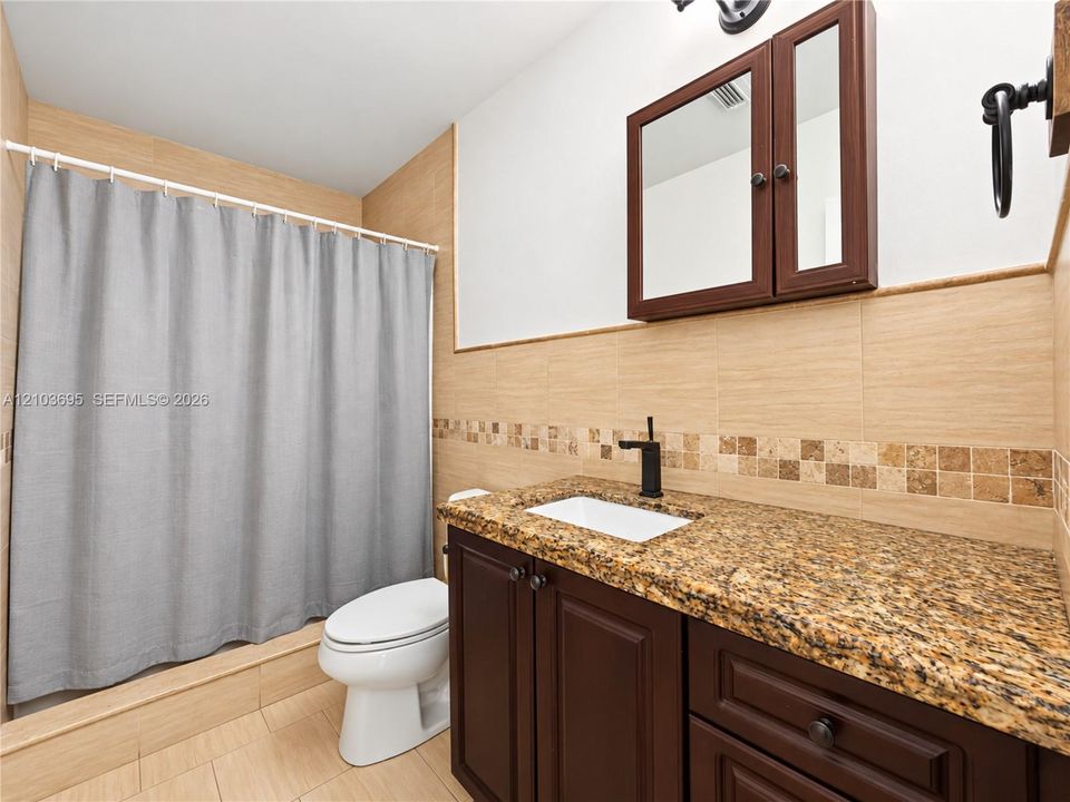 Bathroom featuring stone-surface vanity with rectangular undermount sink, dark wood-finish cabinetry, built-in medicine cabinet, matte black hardware, and tiled wall accents