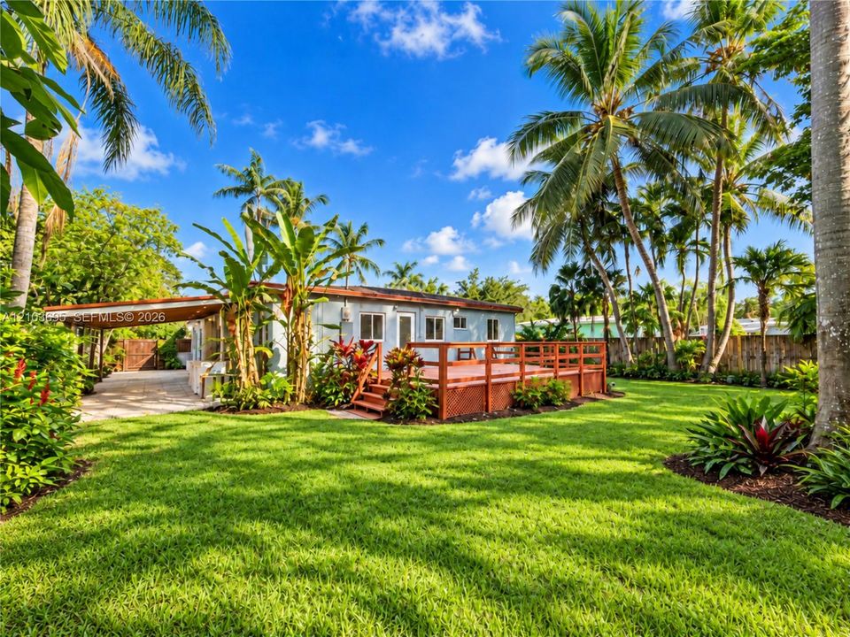 Spacious backyard featuring a wood deck, paved carport, mature palm trees, and extensive landscaping