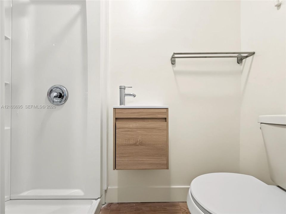 Floating wood-finish vanity with a white countertop and single-handle faucet, integrated shower stall with chrome fixtures, and a wall-mounted metal towel rack