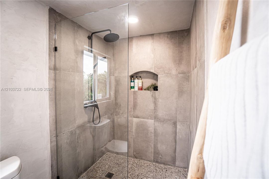 Walk-in shower featuring a glass enclosure, matte black rainfall shower head, pebble-stone flooring, large-format grey tile walls, and a built-in recessed niche