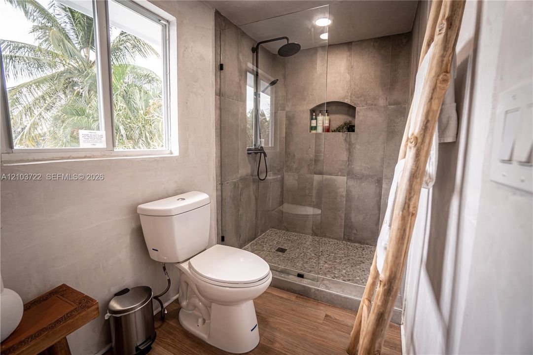 Walk-in shower featuring large-format tile surround, pebble flooring, built-in wall niche, and matte black rainfall shower fixture with handheld attachment, complemented by wood-finish flooring