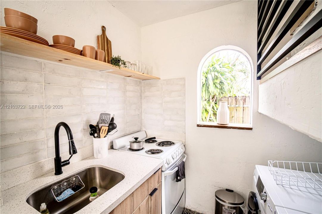 Kitchen featuring a light-toned countertop, undermount stainless steel sink, matte black gooseneck faucet, light-toned wood cabinetry, and a subway-style tile backsplash with an arched window