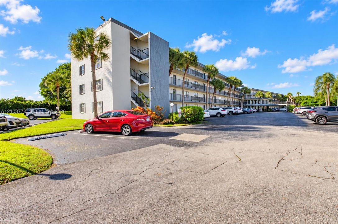Multi-story building featuring exterior balconies, mature palm trees, and a paved parking area