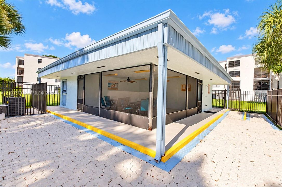 Enclosed structure featuring large window panels and a light-colored exterior, surrounded by a paved patio with a metal fence